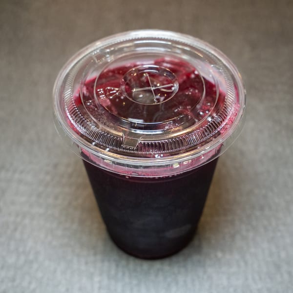 A dark berry smoothie in a clear plastic cup with a lid, on a gray surface.
