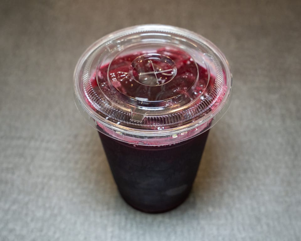 A dark berry smoothie in a clear plastic cup with a lid, on a gray surface.