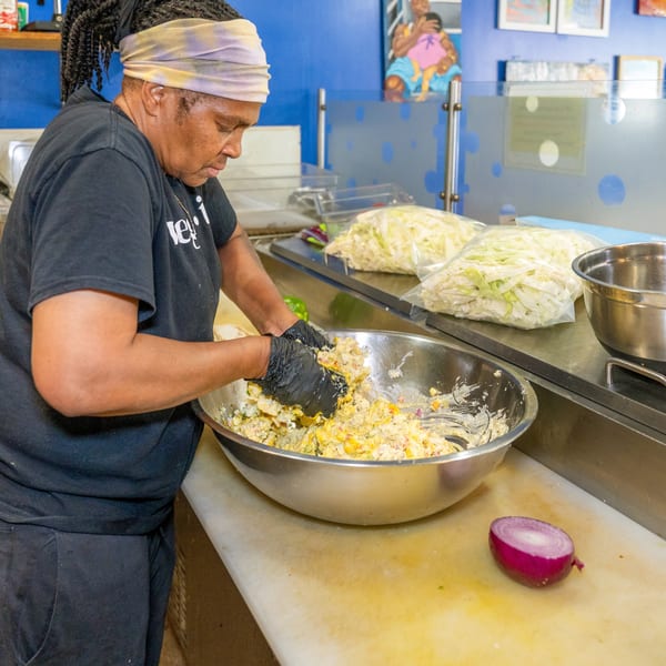 A chef in black gloves mixes a large bowl of potato salad on a kitchen counter.